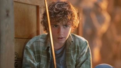 A person with curly hair holds a bow, looking intently forward. They're wearing a plaid shirt and sitting near a wooden box, with a softly lit background.