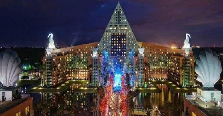 Night view of a large hotel with a pyramid-shaped roof, illuminated with colorful lights. Two large swan statues sit atop shell-like structures on either side. A reflection of the building is visible on the water in front.