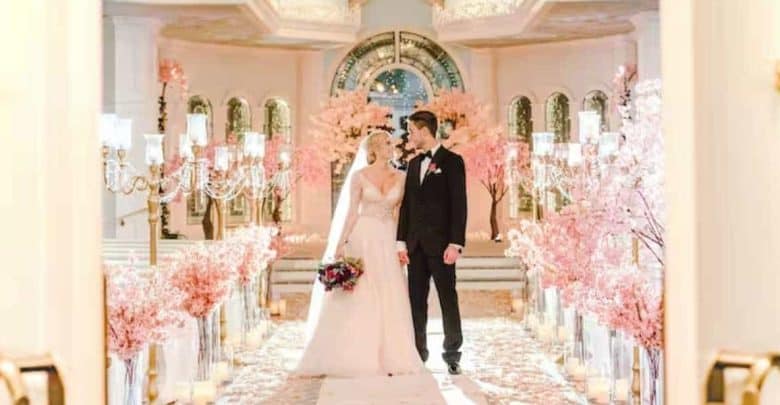 A bride and groom stand together in an elegantly decorated wedding venue. They are surrounded by pink blossom trees and white chandeliers lining the aisle. A large arched window with stained glass is in the background.