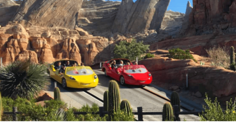 Two colorful convertible cars styled like cartoon characters with eyes on windshields zoom through Radiator Springs Racers in a desert-themed amusement park, weaving past rocky cliffs and cacti under a clear blue sky.