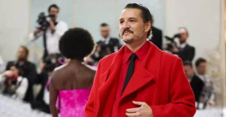 A man in a bright red coat and black tie stands on a red carpet, smiling, with photographers and another attendee in a pink outfit in the background.