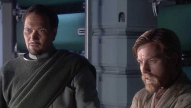 Two men, one with a trimmed beard and dark hair in a gray outfit, and another with short light brown hair and a beard in tan robes, stand indoors beside a metallic wall, both appearing serious and thoughtful.