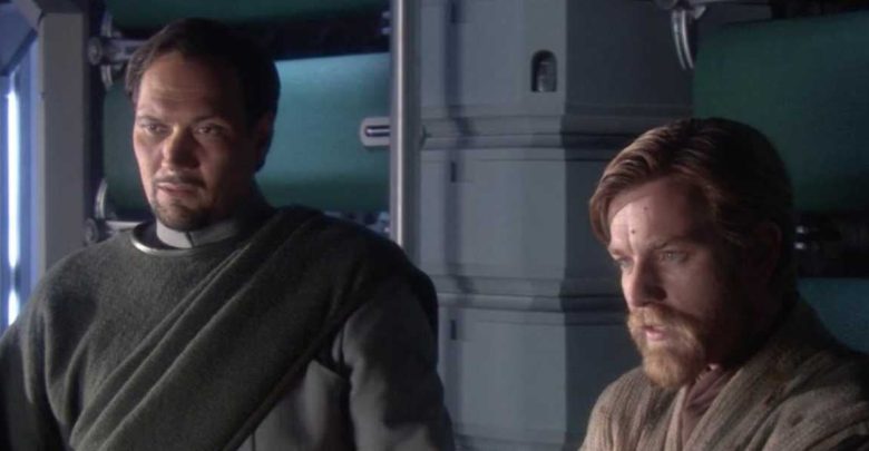Two men, one with a trimmed beard and dark hair in a gray outfit, and another with short light brown hair and a beard in tan robes, stand indoors beside a metallic wall, both appearing serious and thoughtful.