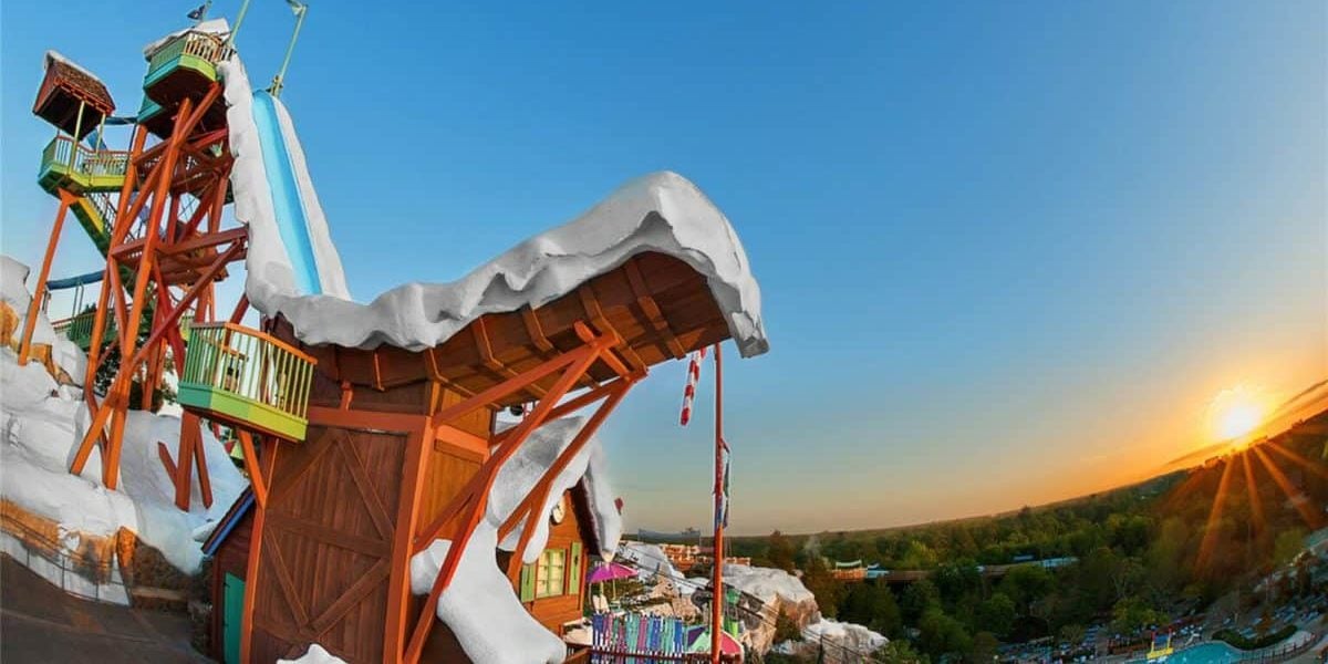 Snow-covered wooden structures with colorful accents rise against a clear blue sky. A tall water slide tower is visible, with a sunset over a green landscape in the background.