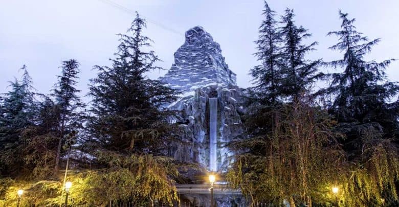 A snowy, rocky mountain peak rises behind tall evergreen trees at dusk, with a waterfall cascading down the center and glowing street lamps illuminating the foreground.