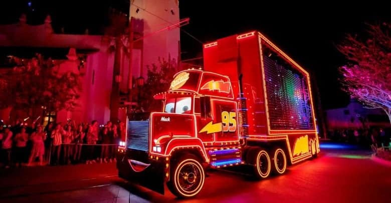 A brightly lit parade float shaped like Mack the truck from the Cars movie, outlined in colorful neon lights, drives past a crowd of spectators at night.