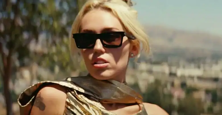 A woman with blonde hair in a messy updo wears black sunglasses, a shiny gold top, and stud earrings, posing outdoors with blurred trees and buildings in the background.