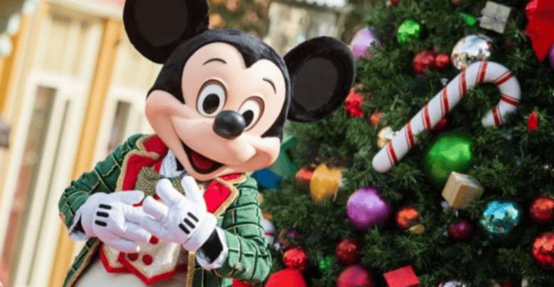 Mickey Mouse, dressed in festive holiday attire, stands smiling in front of a decorated Christmas tree at Christmas Disney World, adorned with ornaments, presents, and a large candy cane.