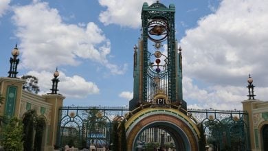 Entrance archway to Universal’s Epic Universe theme park, featuring ornate gold and teal designs, decorative orbs, a large clock tower, and a blue sky with scattered clouds in the background.