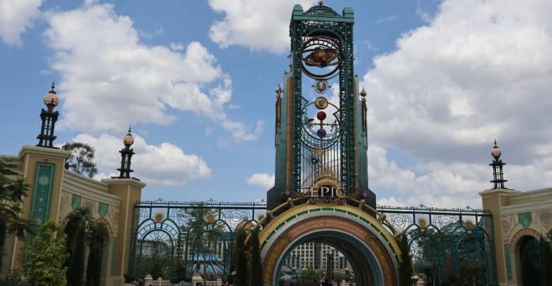 Entrance archway to Universal’s Epic Universe theme park, featuring ornate gold and teal designs, decorative orbs, a large clock tower, and a blue sky with scattered clouds in the background.