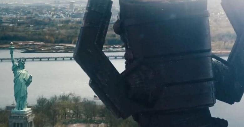 A large robot stands near the Statue of Liberty, towering over the monument, with a river and city visible in the background under a cloudy sky.