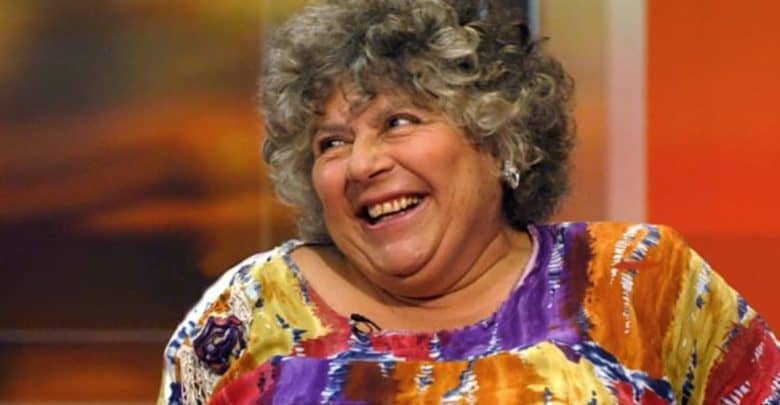 A woman with curly gray hair smiles warmly, wearing a colorful, patterned top. She appears to be sitting indoors, possibly during an interview, with a blurred orange and yellow background behind her.