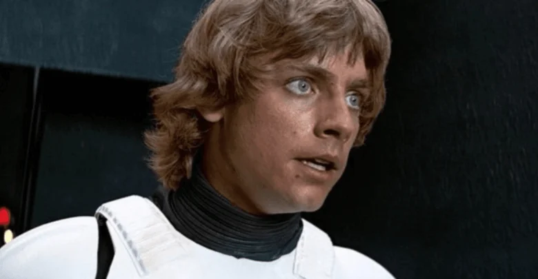 A young man with shaggy blond hair and blue eyes wears a white futuristic armor suit and looks intently ahead, appearing concerned or surprised. The background is dark and metallic.