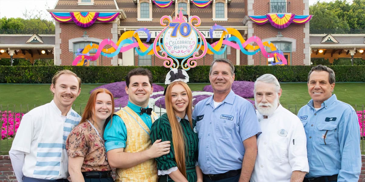 A group of seven people stand smiling in front of a decorated building with colorful banners and a sign reading "70 Celebrated Happy." The scene is festive with flowers and bright decorations.