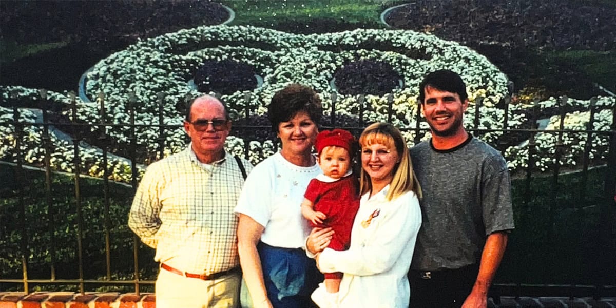 A family of five, including a baby, poses in front of a large flower bed shaped like Mickey Mouse at a theme park, smiling at the camera.
