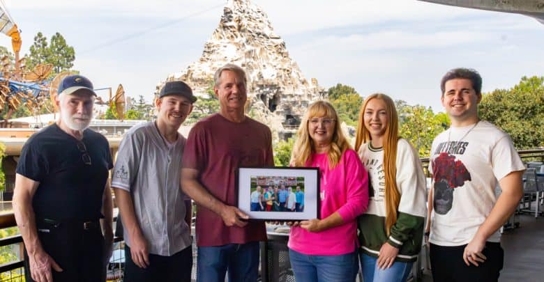 Six people stand together outdoors, smiling at the camera. The group holds a framed photo. A snow-capped mountain ride and trees are visible in the background, suggesting an amusement park setting.