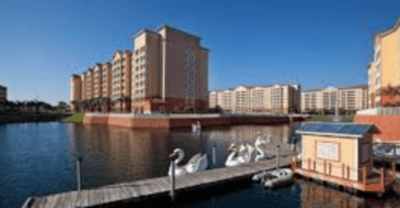 Several tall, modern apartment buildings line the edge of a lake with a dock in the foreground. Swan-shaped pedal boats and a small wooden structure are visible by the dock under a clear blue sky.