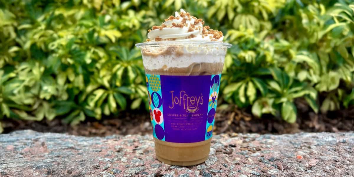 A Joffrey's iced coffee drink topped with whipped cream and crumbled toppings, served in a clear plastic cup with a colorful purple label, sits on a stone surface with green foliage in the background.