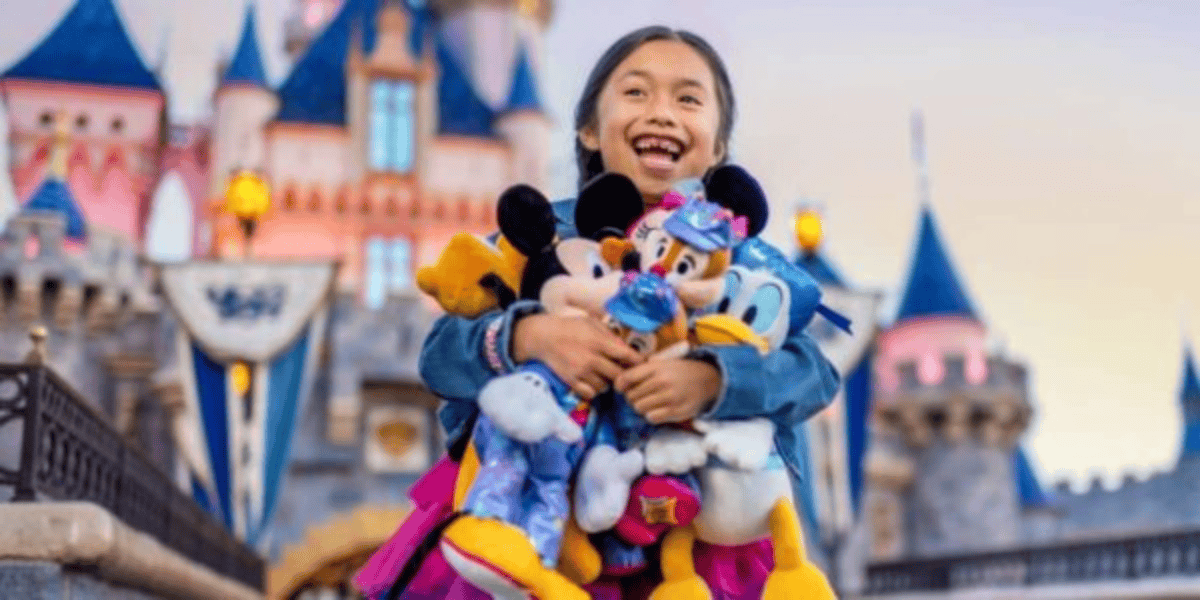 A joyful child holds multiple plush Disney characters, including Mickey, Minnie, and Donald, in front of a colorful fairy-tale castle at a theme park. The background features spires and flags, suggesting a magical atmosphere within Disney World and Disneyland.