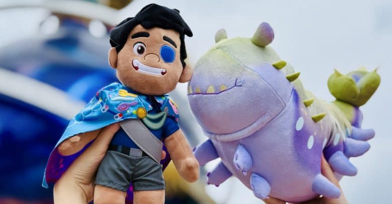 Two hands hold up plush toys: one is a smiling boy with black hair, an eye patch, and a colorful cape; the other is a friendly-looking, pastel purple-and-green creature with small legs and a finned tail.
