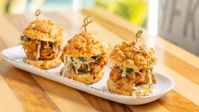 Three fried chicken sliders with coleslaw are served on a white oval plate, each held together with a wooden pick, on a wooden table.