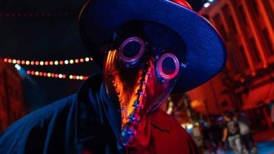 A person wearing a dark wide-brimmed hat, a long black coat, and a metallic plague doctor mask with round goggles and a beak, stands under string lights in a city street at night with red and blue lighting.