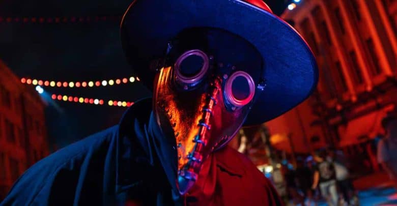 A person wearing a dark wide-brimmed hat, a long black coat, and a metallic plague doctor mask with round goggles and a beak, stands under string lights in a city street at night with red and blue lighting.