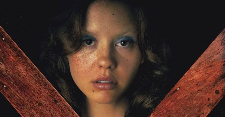 A young woman with wavy brown hair and blue eyeshadow stares ahead. Two large, blood-stained wooden planks form an X in front of her against a dark background.