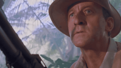 A man in a safari hat and khaki clothing looks ahead intensely, holding a large gun, with lush jungle foliage and sunlight in the background.