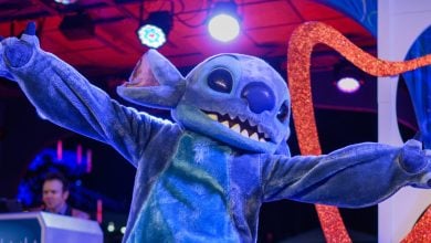 A person in a large, blue, furry Stitch costume from the movie "Lilo & Stitch" poses with arms wide open under colorful stage lights at an indoor event.