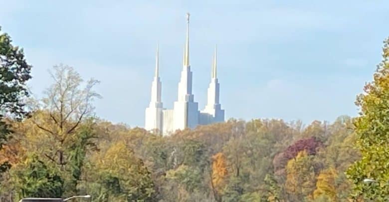 A white temple with five tall spires rises above a forest of autumn trees with colorful foliage under a clear sky.