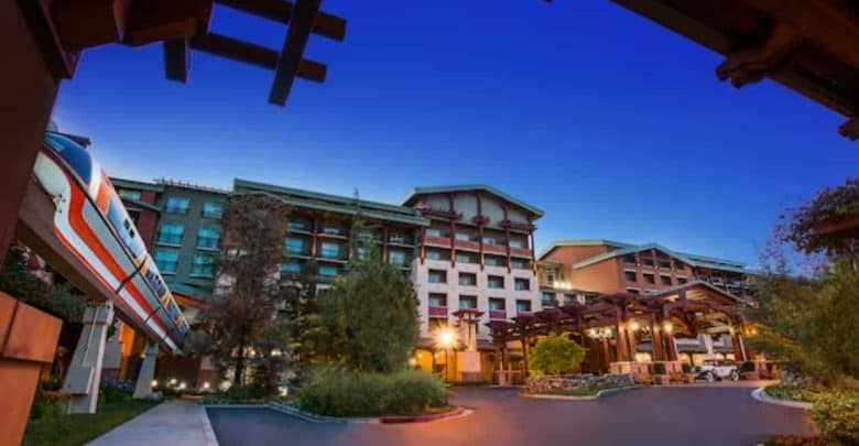 A sleek monorail passes by the entrance of a large, well-lit resort hotel with wooden architectural details and lush greenery at dusk, under a clear blue sky.