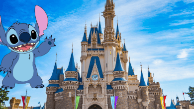 Cartoon character Stitch waves in front of a large Disney-style castle with blue spires, under a bright blue sky with some clouds. Colorful banners hang on the castle.