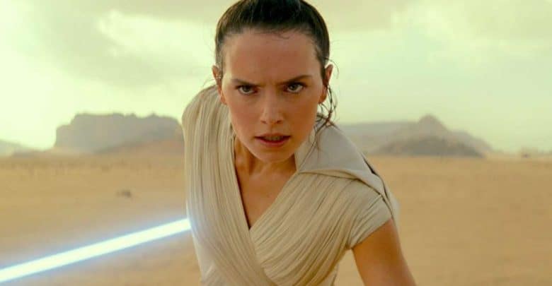 A woman with brown hair tied back holds a lit blue lightsaber in front of her, standing in a desert landscape with rocky formations in the background. She looks determined and focused.
