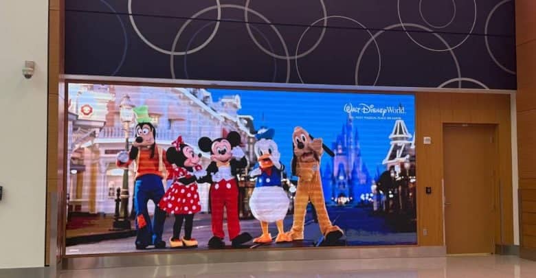 A digital display shows Disney characters Goofy, Minnie Mouse, Mickey Mouse, Donald Duck, and Pluto smiling and waving, with Cinderella Castle and Main Street, U.S.A. in the background inside Orlando International Airport