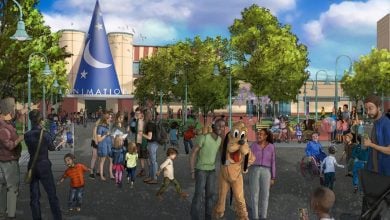 A lively theme park scene with families, children, and a person in a Pluto costume. Trees, lampposts, and a building with a large blue wizard hat labeled “ANIMATION” are in the background under a partly cloudy sky.