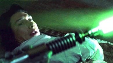 A person lying on their side looks back in fear as a glowing green lightsaber is ignited near their face, illuminating the scene with green light.