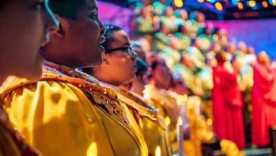 A choir dressed in yellow and red robes sings on stage, holding candles, with colorful lights and a blurred audience in the background.