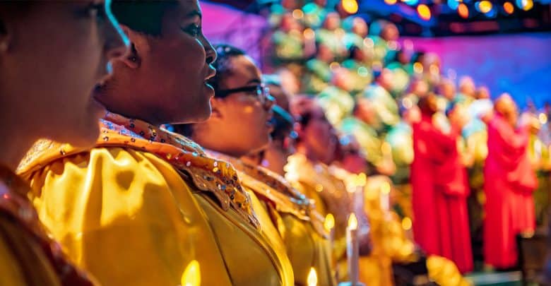 A choir dressed in yellow and red robes sings on stage, holding candles, with colorful lights and a blurred audience in the background.