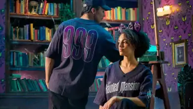 Two people in a room with bookshelves and purple wallpaper decorated with green eyes. One person wears a "999" jersey and cap, the other sits on a stool in a "Haunted Mansion" jersey with cat ear headband.