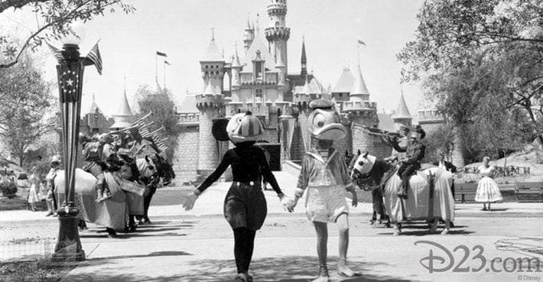 Two people in vintage Donald Duck costumes walk hand-in-hand in front of Sleeping Beauty Castle at Disneyland, with band members on horseback and park guests in the background. The image is in black and white.