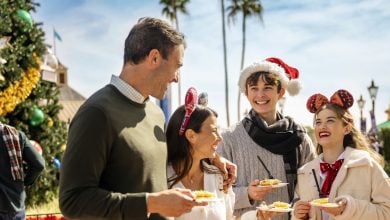 A group of four people, two men and two women, smile and enjoy food outdoors near a decorated Christmas tree. One person wears a Santa hat, another wears festive Mickey ears. Palm trees and clear skies are in the background.