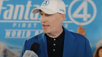Kevin Feige, in a blue suit and white "Fantastic Four" cap, talks about Fantastic 4 as he speaks into a microphone at the "Fantastic Four: First Family World Premiere," standing before a themed blue backdrop.