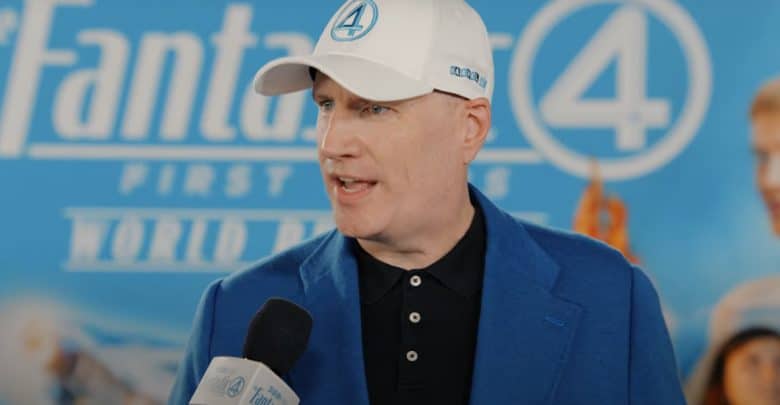 Kevin Feige, in a blue suit and white "Fantastic Four" cap, talks about Fantastic 4 as he speaks into a microphone at the "Fantastic Four: First Family World Premiere," standing before a themed blue backdrop.