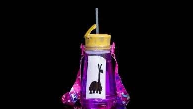 A purple water bottle with a yellow lid and straw, featuring a cartoon giraffe sticker and a pink strap with colorful patterns, is set against a black background.