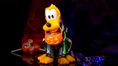 A plastic Pluto figurine dressed in a skeleton costume holds a pumpkin-shaped Halloween bucket filled with candy. Colorful lollipops are visible in the background, set against a dark backdrop.