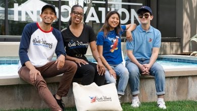 Four smiling people sit together by a fountain outside. They wear casual clothing featuring "Walt Disney Imagineering" logos and Disney characters. A large cream tote bag with a matching logo is placed in front of them.