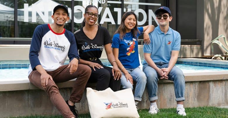 Four smiling people sit together by a fountain outside. They wear casual clothing featuring "Walt Disney Imagineering" logos and Disney characters. A large cream tote bag with a matching logo is placed in front of them.