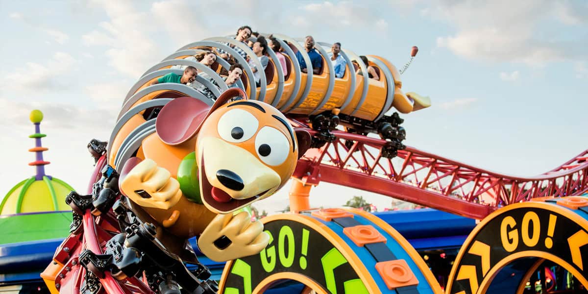 People ride a colorful roller coaster shaped like Slinky Dog from Toy Story, curving on bright red tracks with “GO!” signs under a blue sky—one of the Walt Disney World rides to avoid if you dislike thrilling twists at amusement parks.