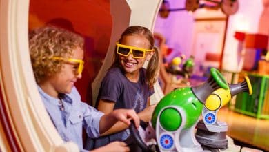 Two smiling children wearing yellow 3D glasses play with toy cannons at an indoor Disney's Hollywood Studios attraction, surrounded by colorful decorations and a playful atmosphere.